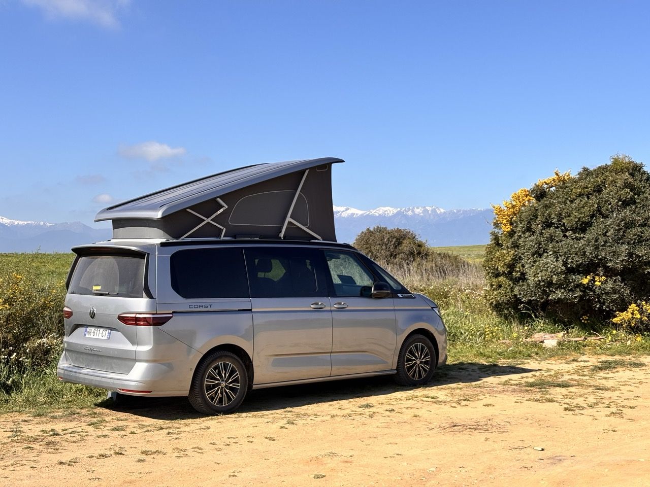Le Volkswagen California Coast toit relevable déployé, garé sur une piste de terre bordée d'ajoncs en fleur, face à des montagnes enneigées